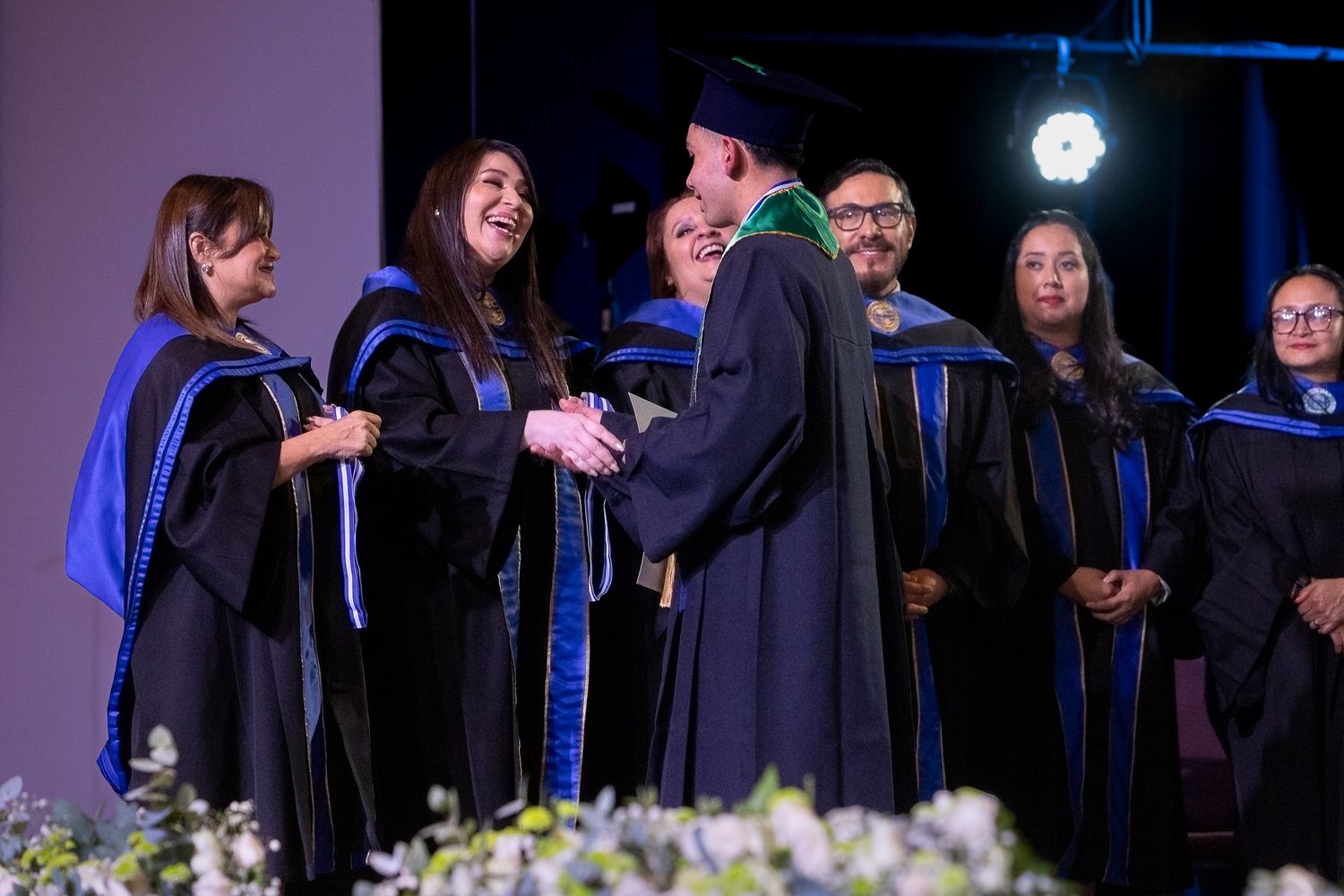 Graduates in black caps and gowns receive diplomas during a formal commencement ceremony with blue stage lighting.