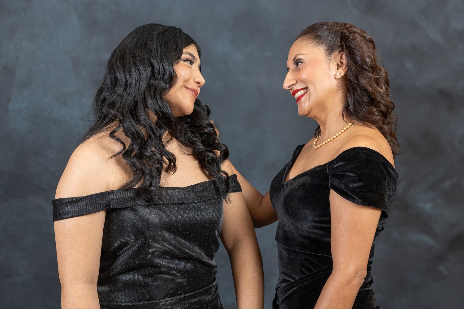 Two women in elegant black off-shoulder dresses share a joyful moment against a dark background.