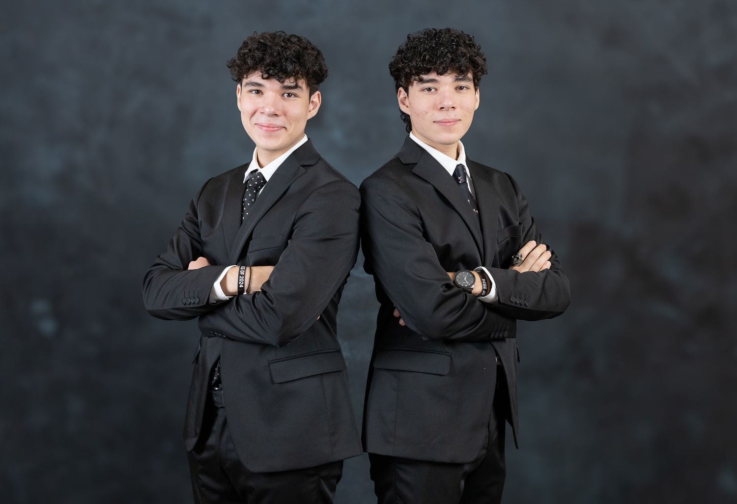 Professional studio portrait of two young men in black suits against a dark background.