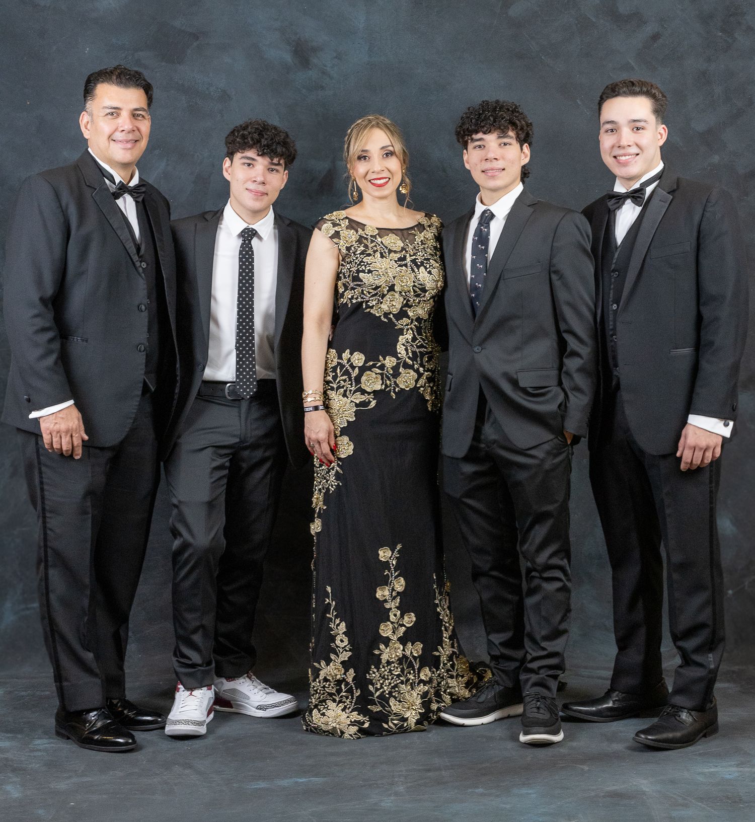 Formal group portrait with people in black suits and one wearing a gold and black evening gown against a dark backdrop.