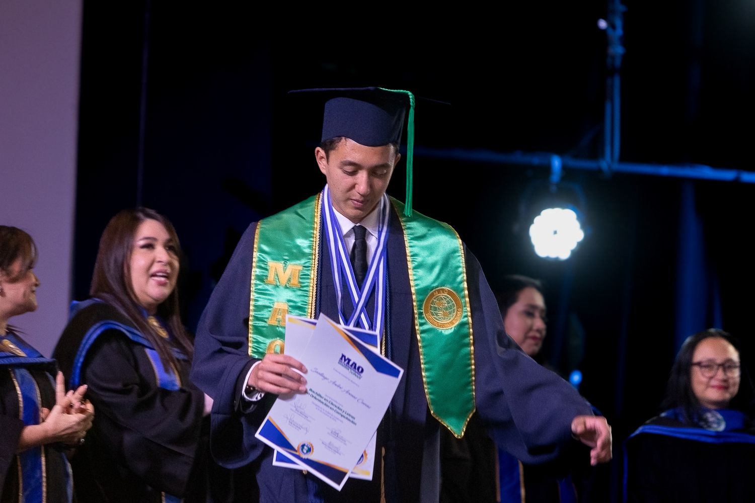 Students in formal attire celebrate with diplomas during graduation ceremony with dark blue stage lighting.