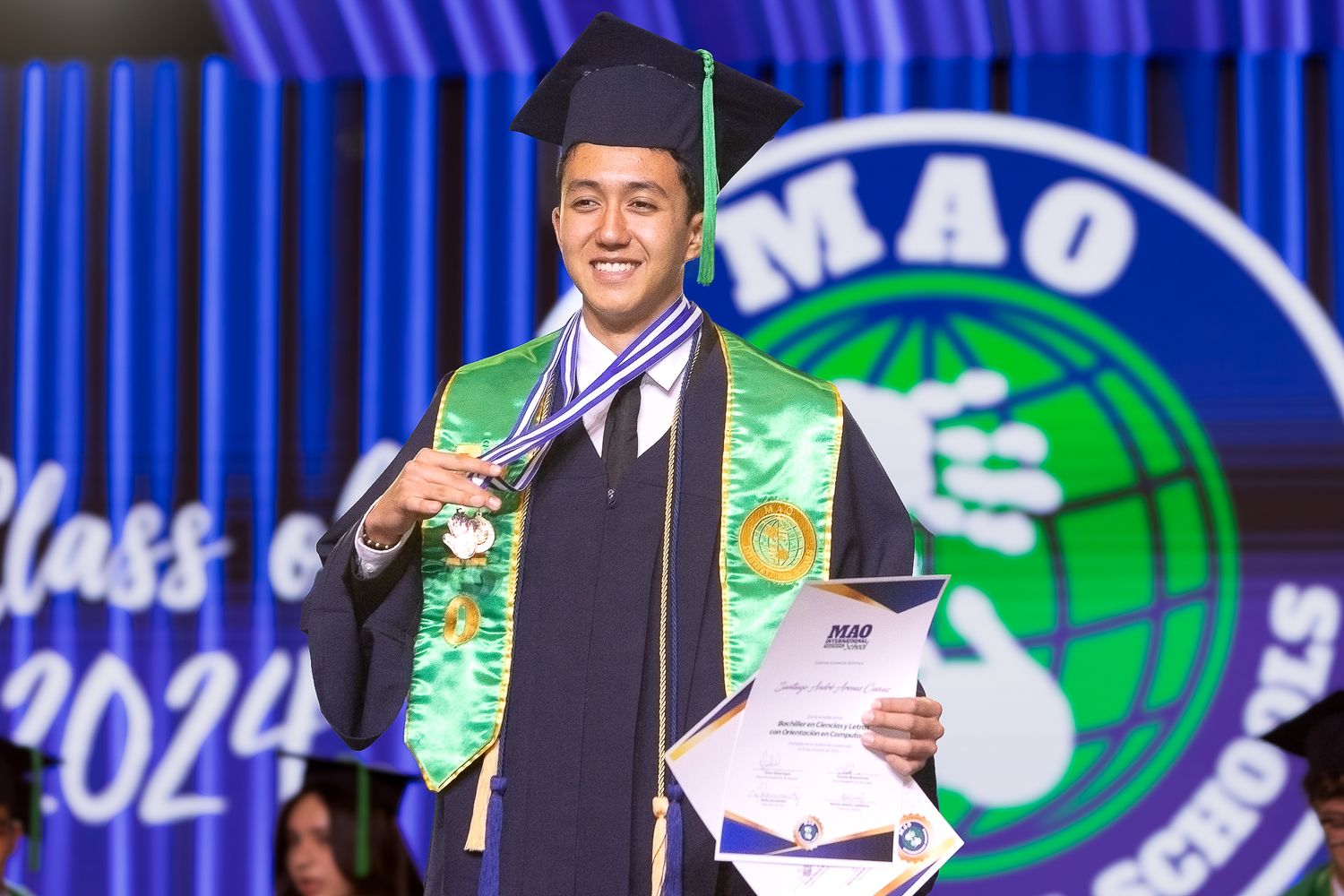 Graduates in green and white academic regalia stand in front of a bright blue illuminated backdrop during a ceremony.
