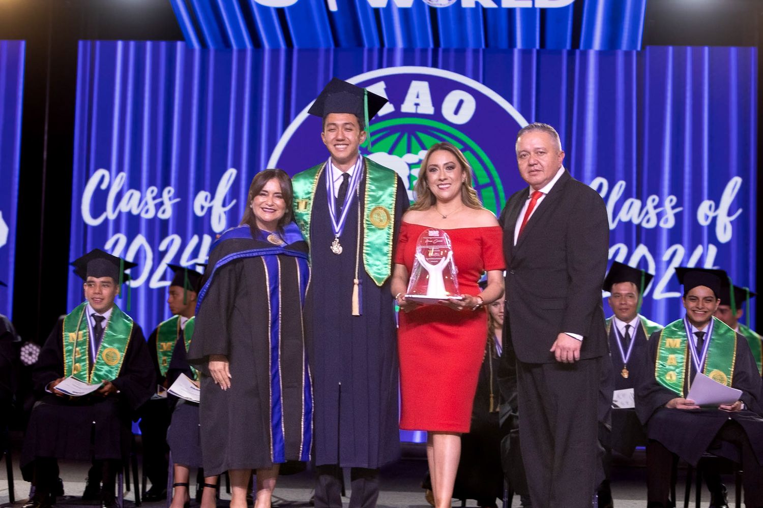 Graduates in blue caps and gowns stand on stage during a commencement ceremony with illuminated blue lighting.