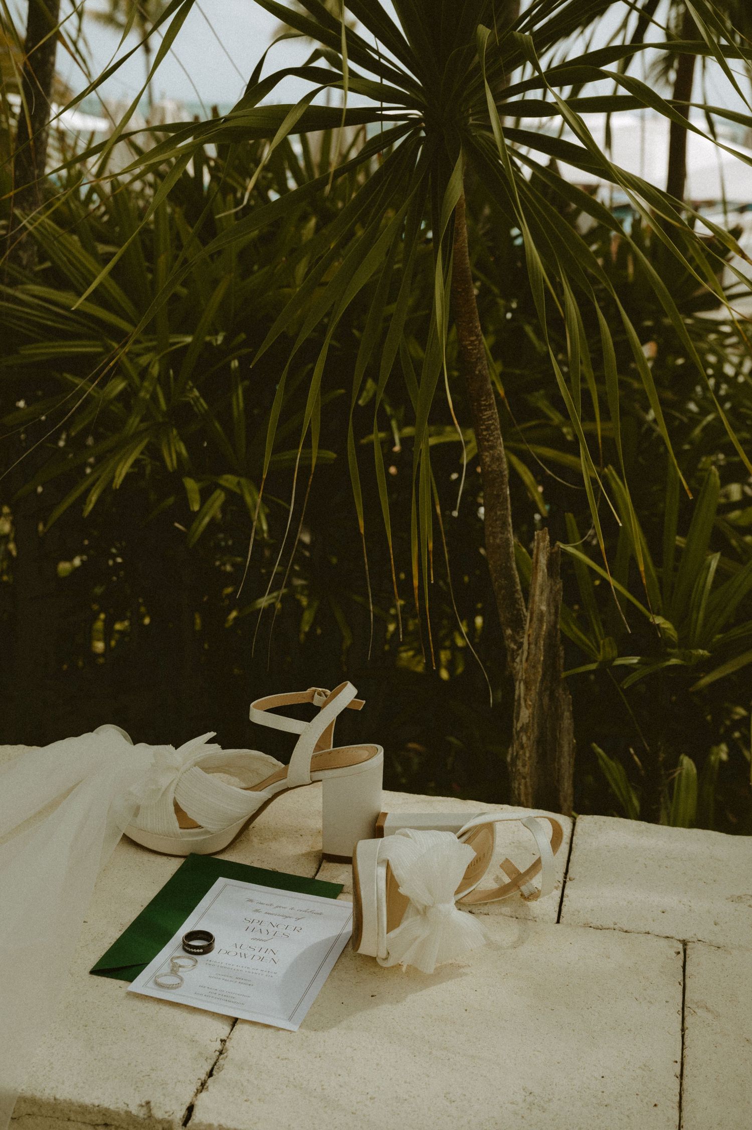 White sandals and a book rest on a concrete surface against tropical palm fronds in moody lighting.