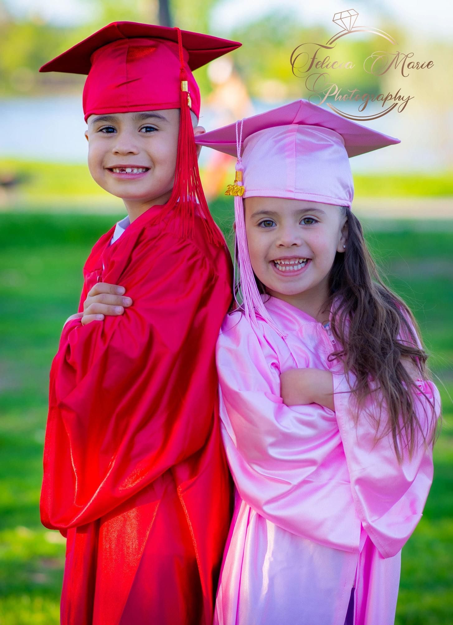 Two young graduates wearing caps and gowns in red and pink stand back-to-back outdoors smiling on their special day.