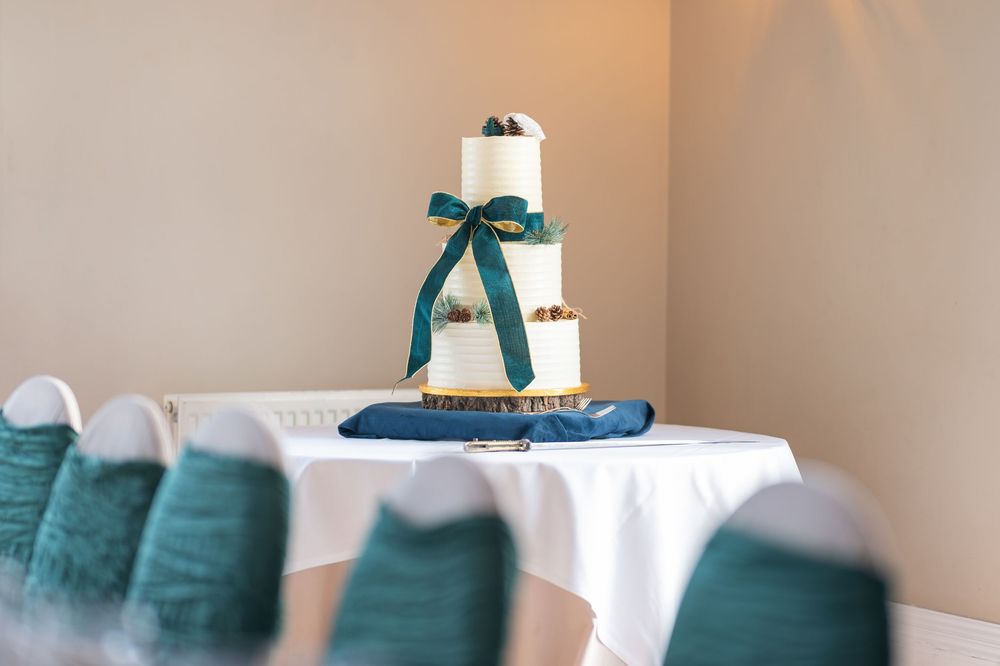 White tiered wedding cake with elegant teal ribbon bow displayed on white tablecloth with matching teal chair covers.