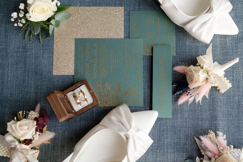 Wedding invitation suite with teal and kraft paper cards surrounded by blush roses and white napkins on blue linen.