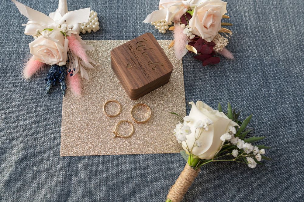 Rustic wedding details including flowers, rings, and wooden decor arranged on gray fabric backdrop.