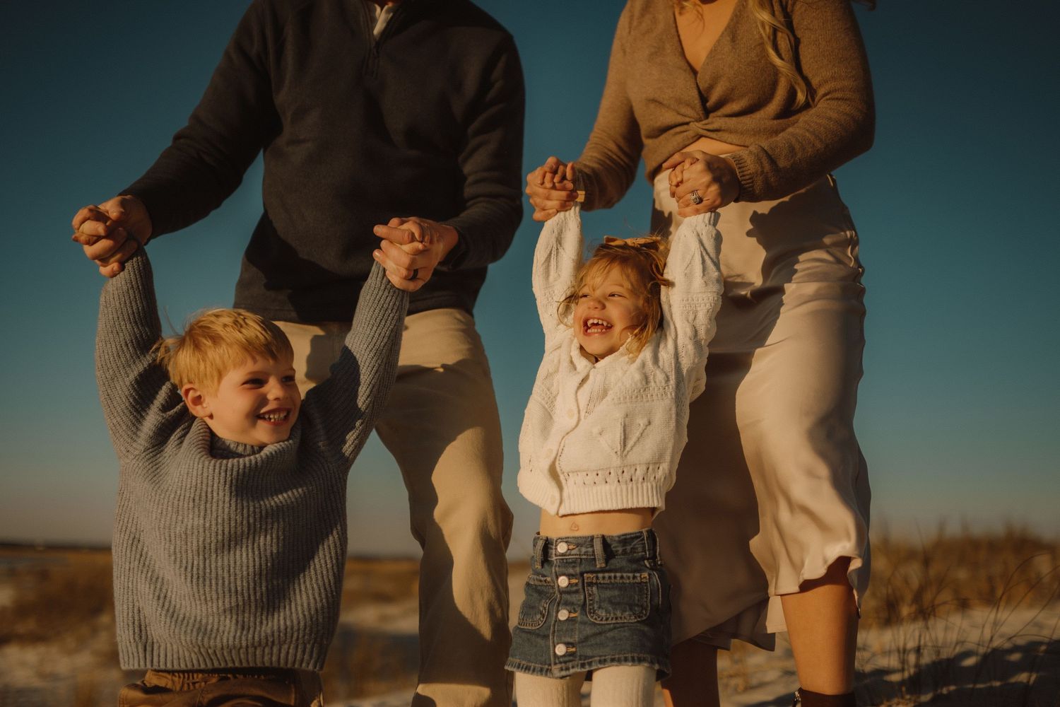 Family and their black dog pose together on a sandy beach at sunset in matching neutral colored outfits.