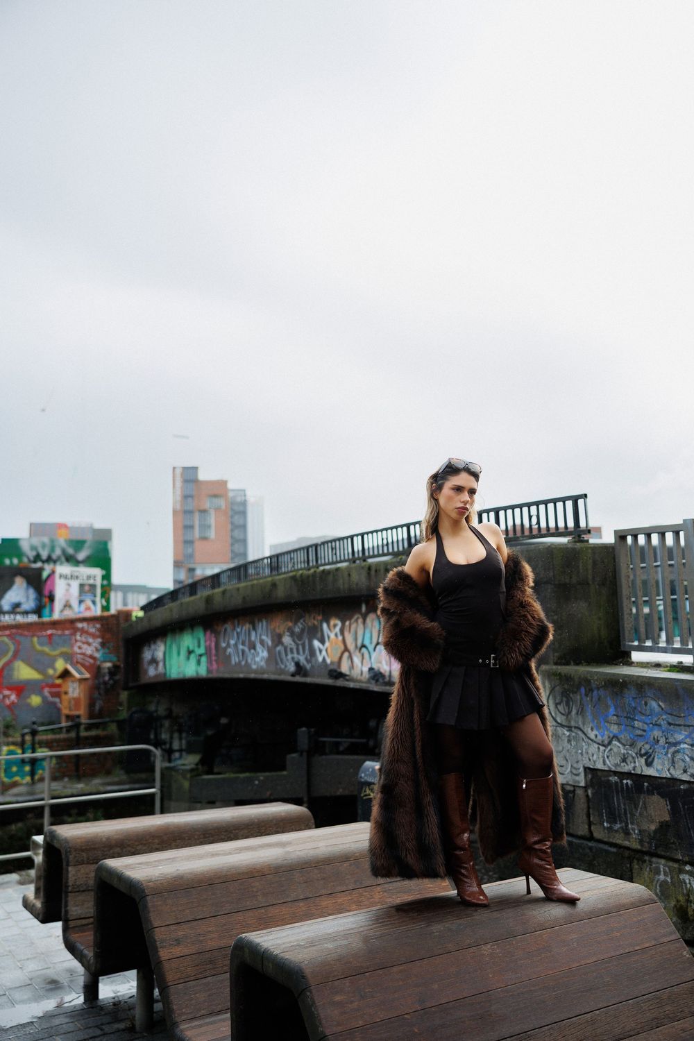 Person in dark clothing poses on wooden bench against urban backdrop with graffiti and metal railings.