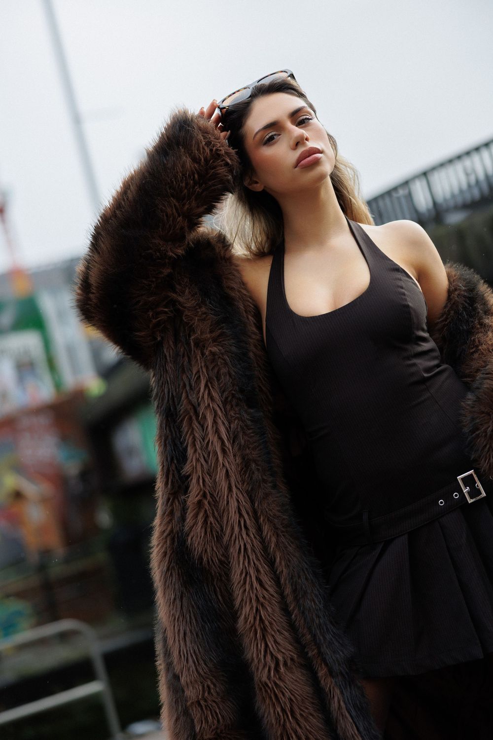 Fashion model wearing black top and brown fur coat poses dramatically against urban backdrop.
