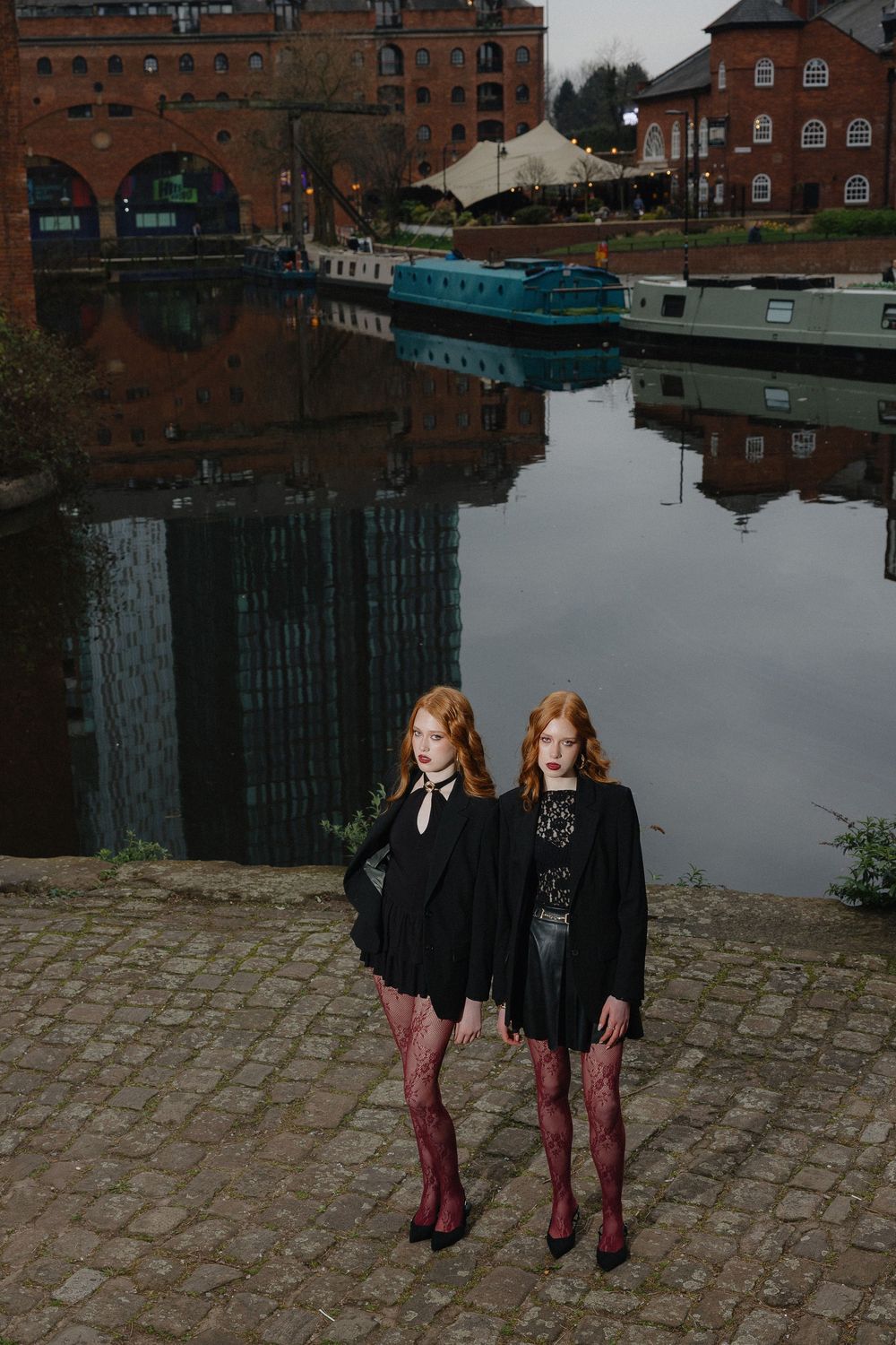 Two figures in dark outfits and burgundy tights stand on cobblestones by a canal with brick buildings and boats in view.