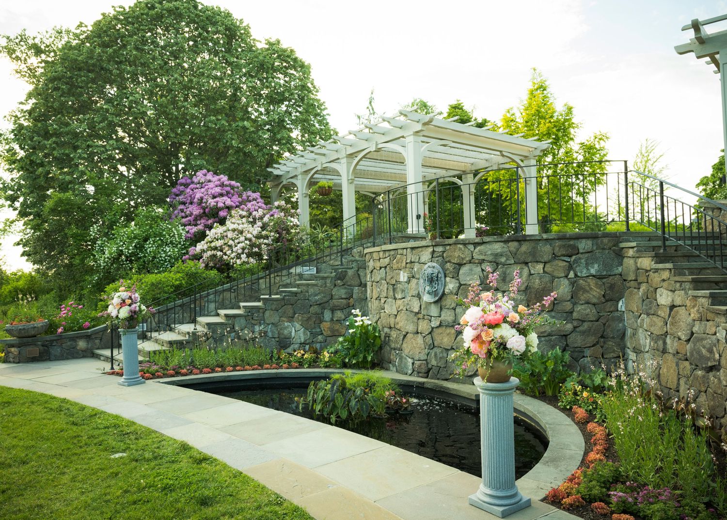 White pergola sits atop a curved stone retaining wall with landscaped garden and walkway in a peaceful park setting.