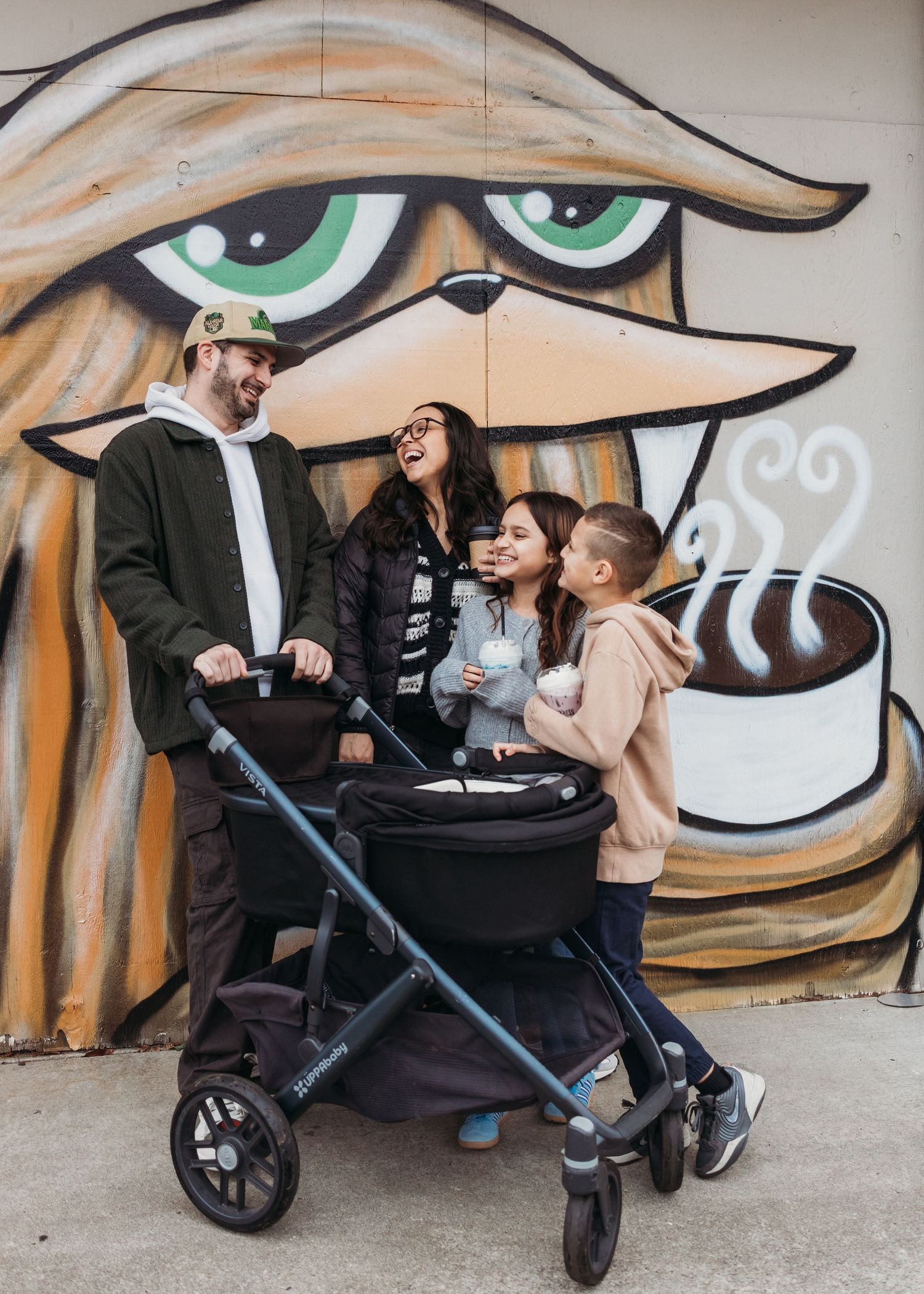 Family sitting together against urban graffiti wall with large cartoon eyes and mustache artwork in background.