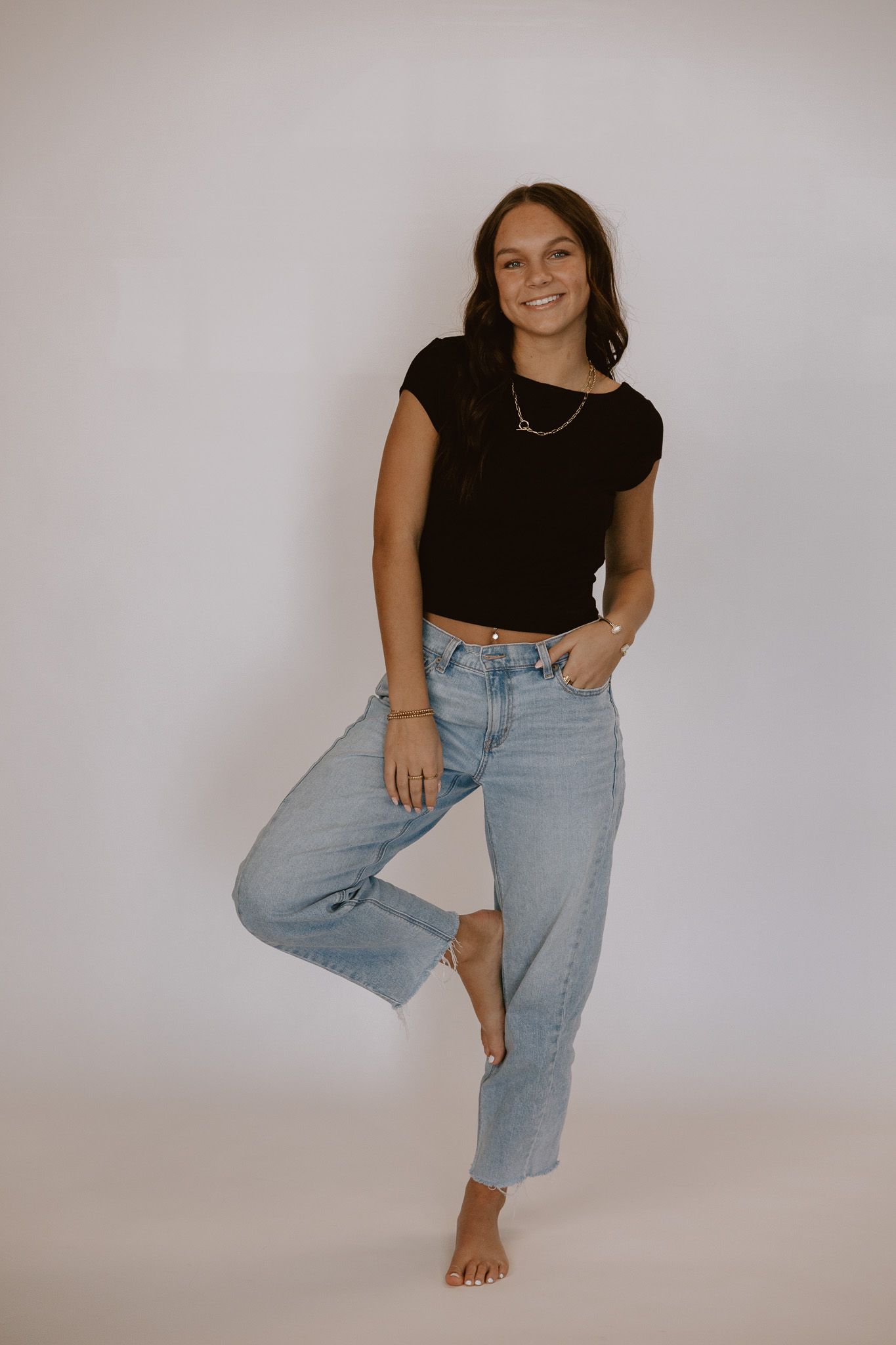 Smiling young woman in black crop top and light wash jeans poses barefoot against white background.