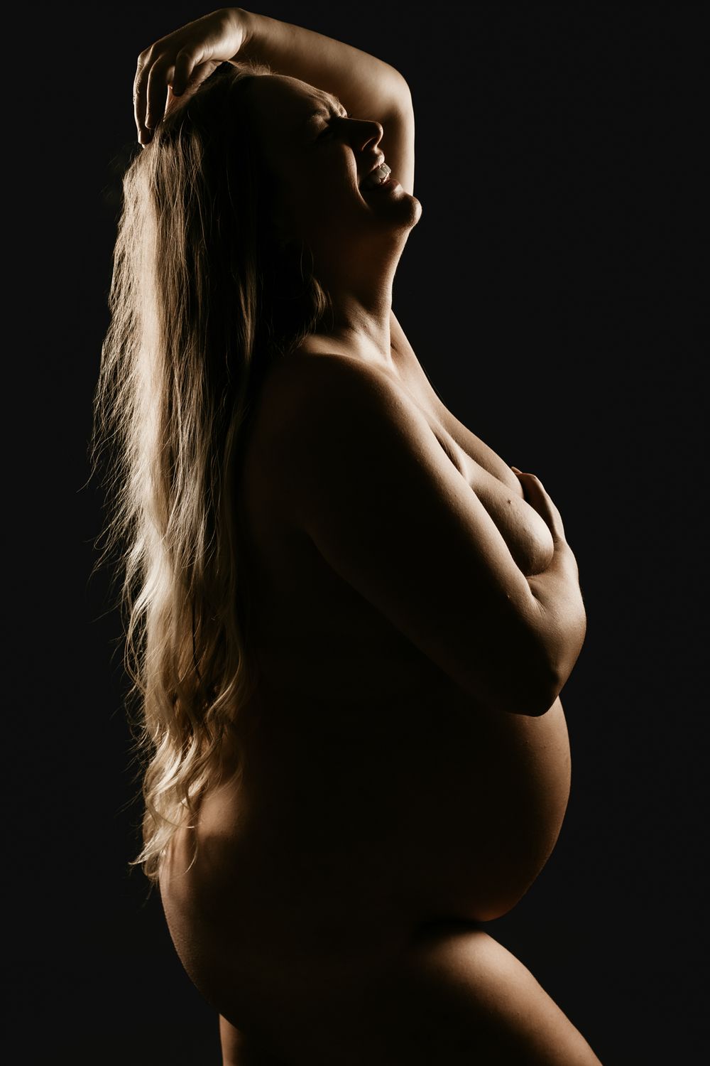 Artistic silhouette of a pregnant figure with long hair against dark background showing dramatic side lighting.