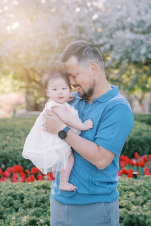 A parent in a blue shirt holds and embraces a small baby wearing white in a garden with red flowers.
