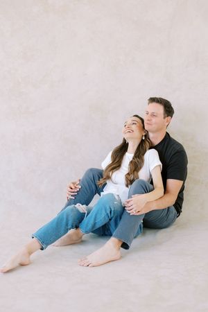 A couple in casual outfits sitting close together on a light colored floor, sharing a candid laughing moment.