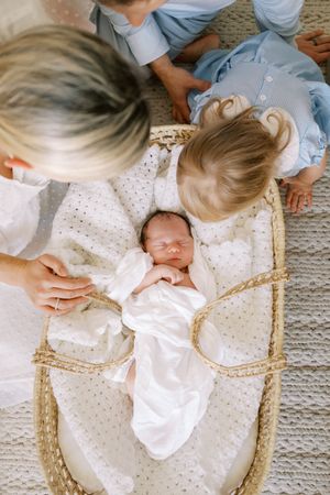 A newborn baby lies in a woven bassinet on a carpet while being tended to by others nearby.