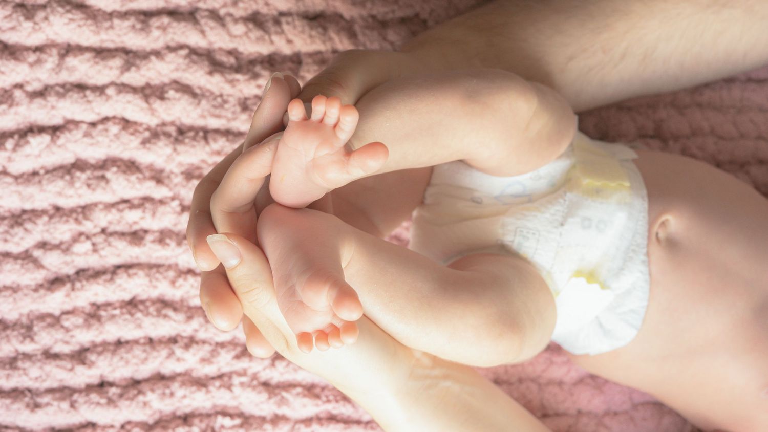 Baby wearing white diaper lies on soft pink knitted blanket with tiny feet and legs visible.