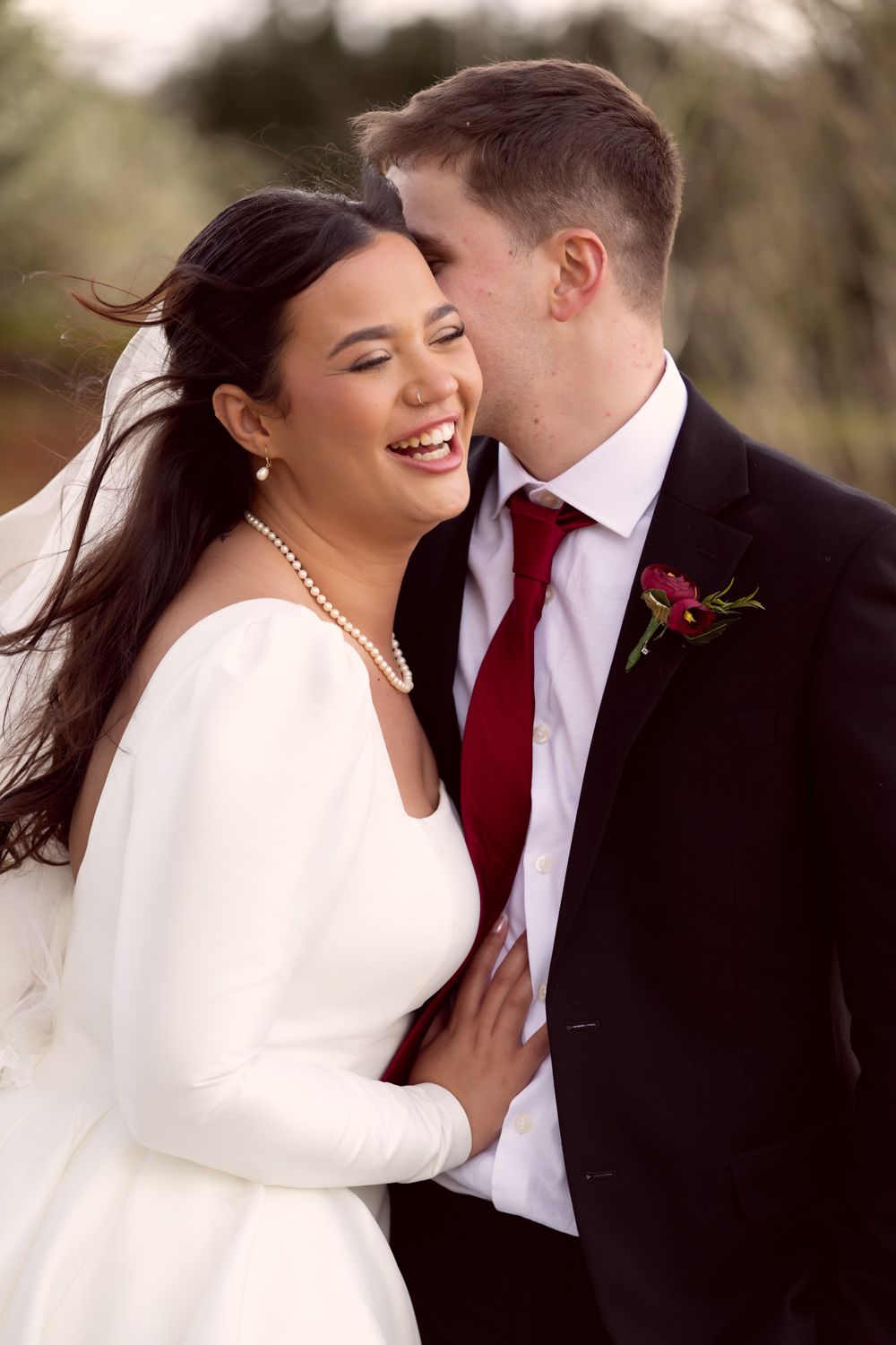 A joyful wedding moment captured outdoors with the bride in a long-sleeved white dress and groom in black suit with red tie.