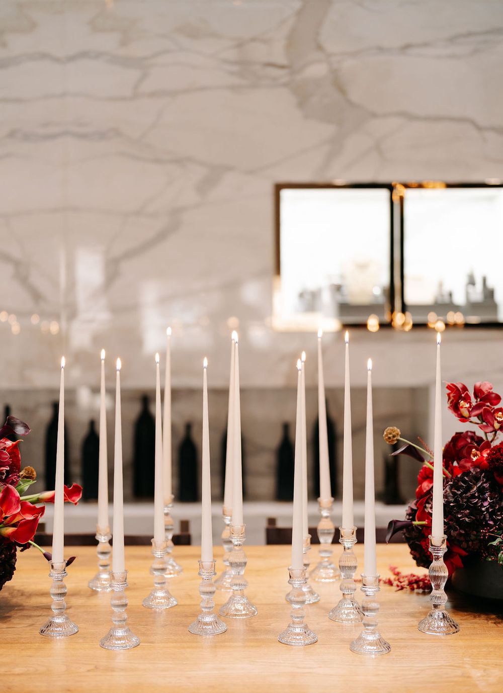 Multiple white candles line a wooden table with red floral decorations and pine cones as elegant holiday decor.