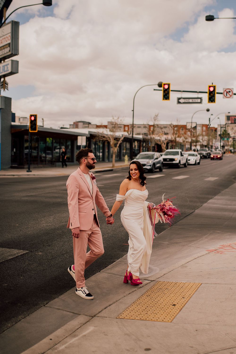 A couple in coordinated pink and white formal attire walk hand in hand down an urban street.