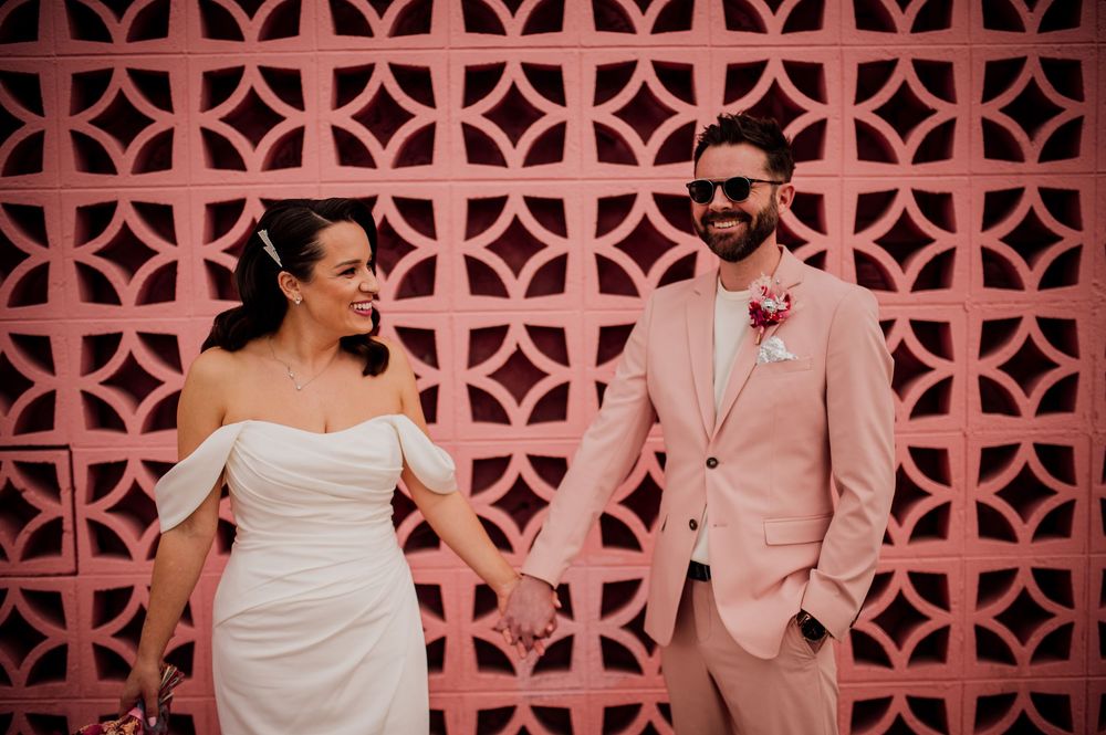 A couple in light pink and white formal attire poses together against a decorative coral-colored lattice wall.