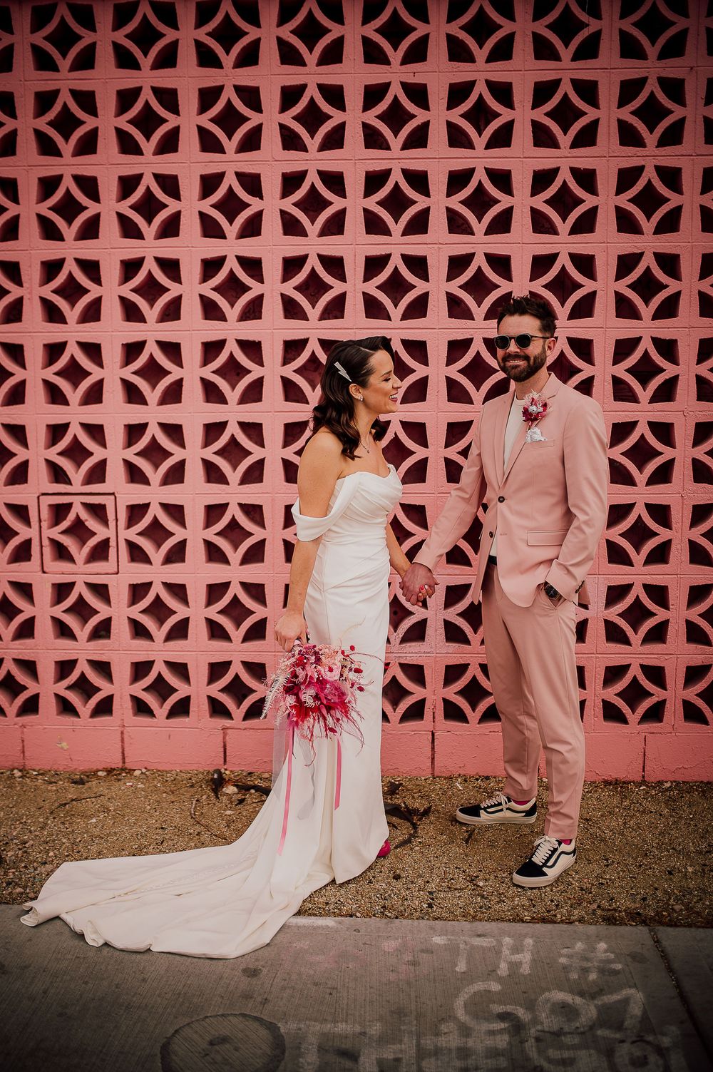 Wedding couple poses against pink geometric breeze block wall with bride in white dress and groom in pink suit.