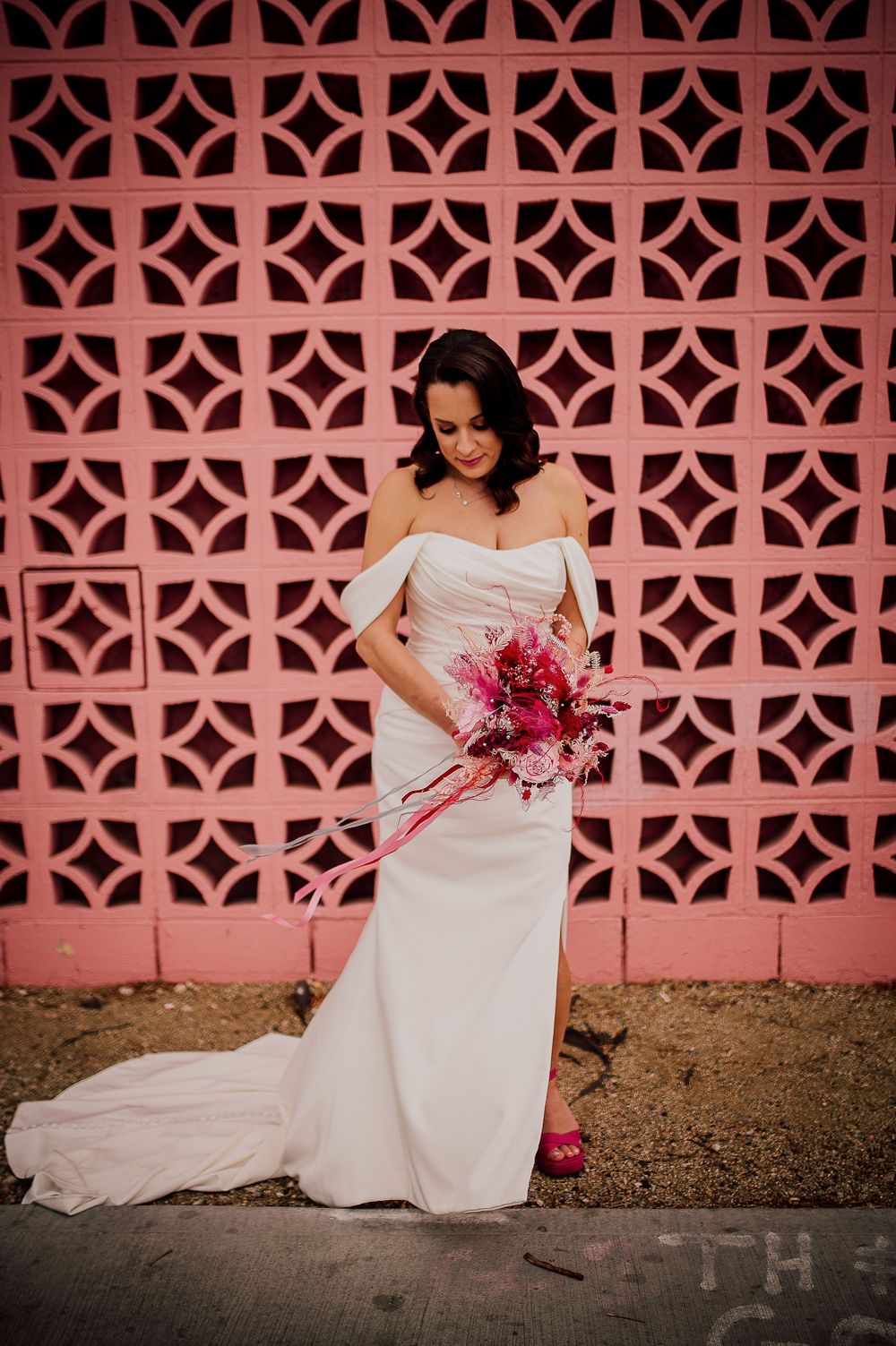 A bride in a white wedding dress poses against a pink decorative wall while holding a vibrant pink bouquet.