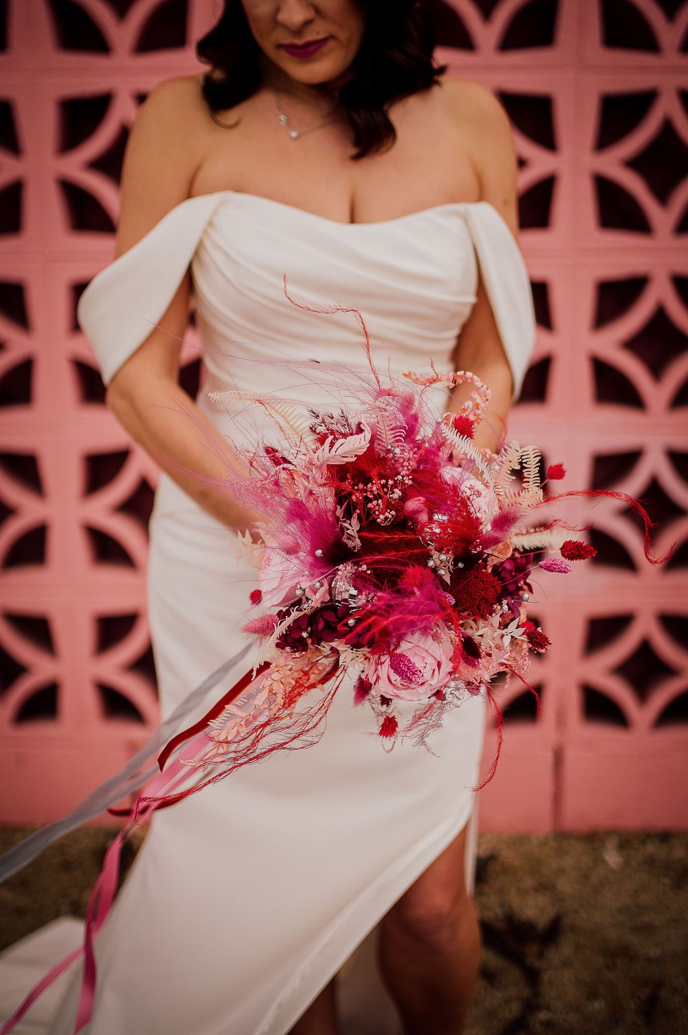 A stunning pink and red bridal bouquet pops against a white wedding dress and coral geometric background.