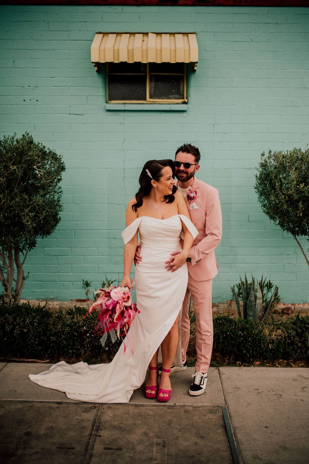 Modern wedding couple poses against mint green wall with pink bouquet and unique style choices including sneakers and pink heels.