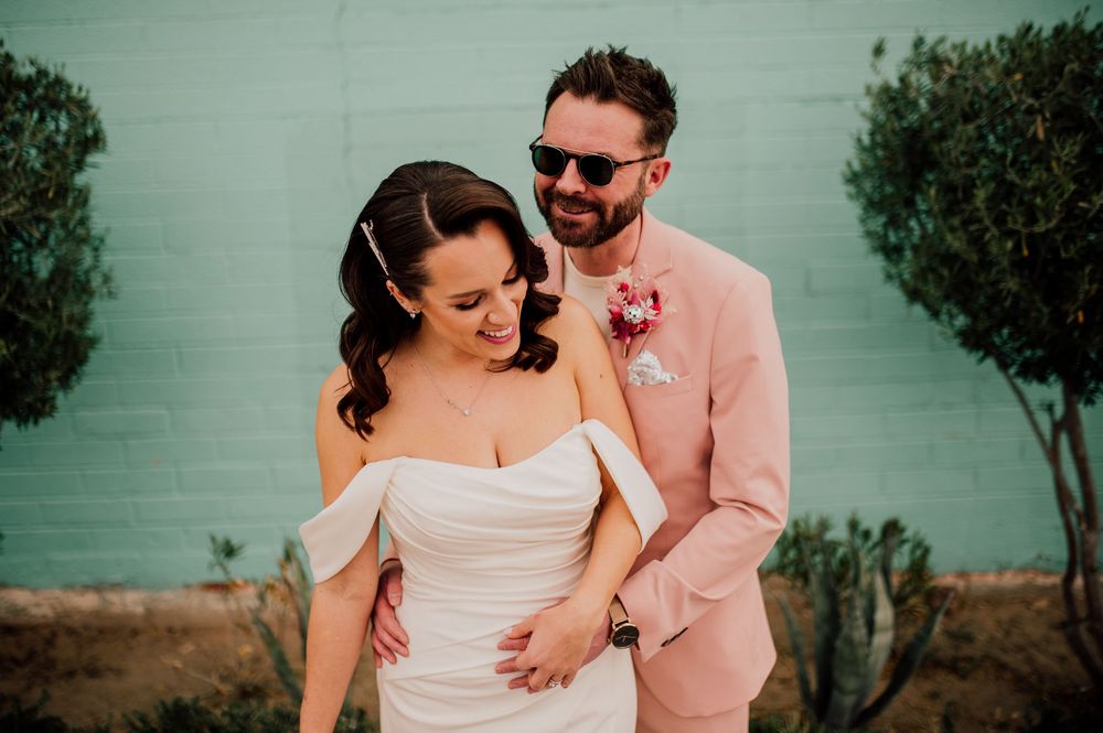 A couple in wedding attire share a candid moment against a mint green wall, with the groom in a pink suit.