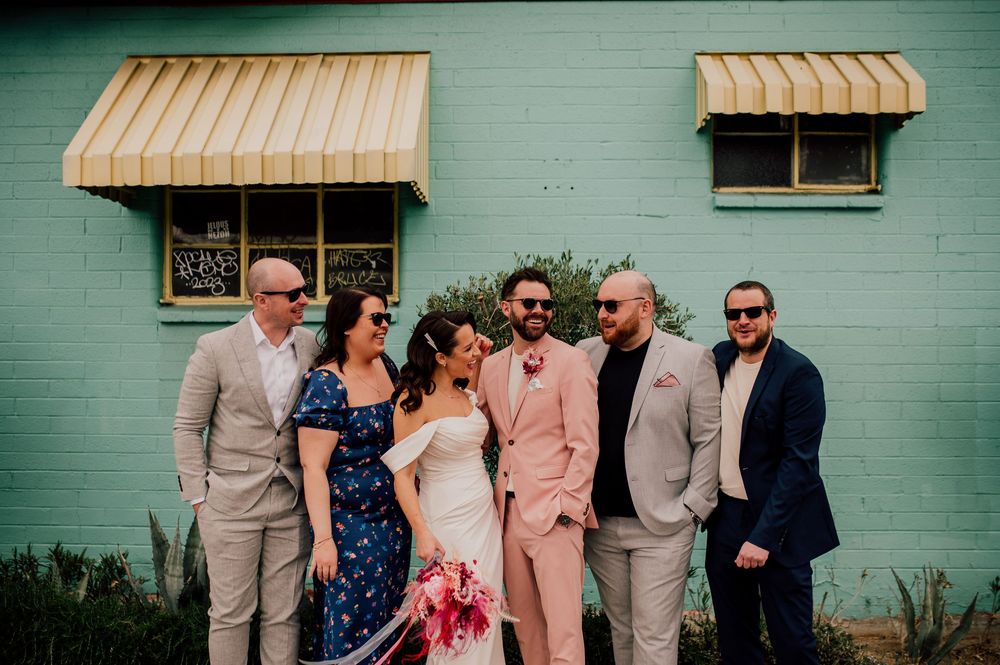 Wedding party in formal attire poses together against mint green wall with striped awnings.