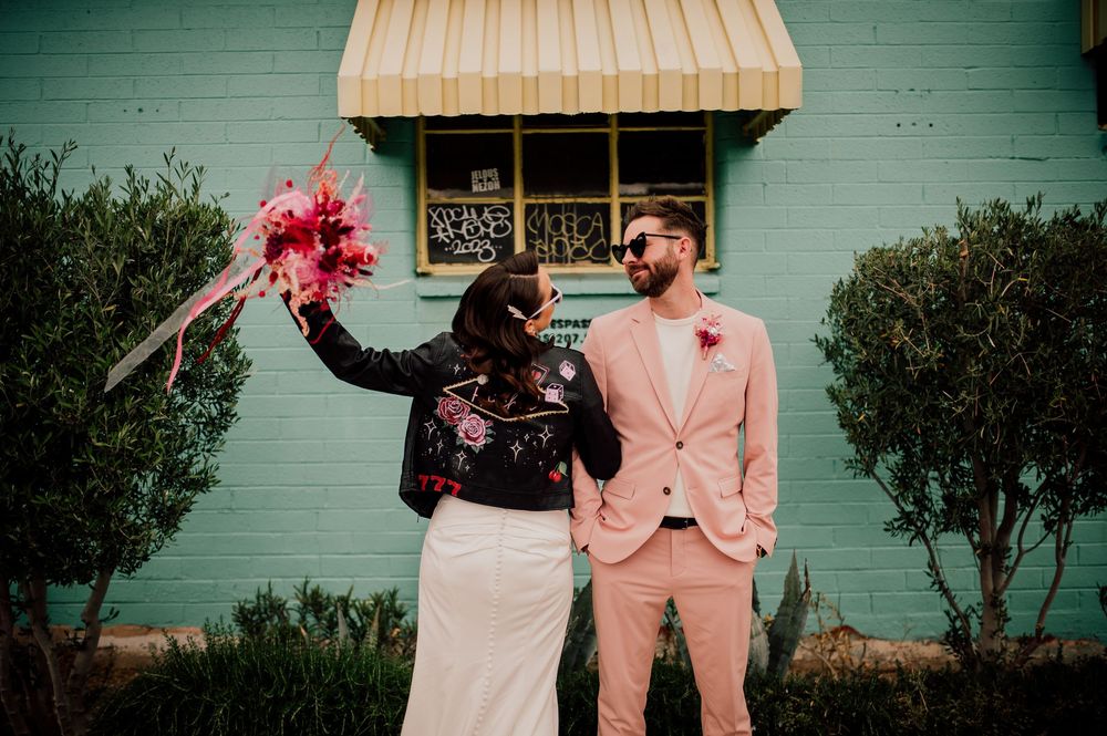 A joyful couple celebrates in front of a mint green wall, with one holding bright pink flowers while wearing a peach suit.