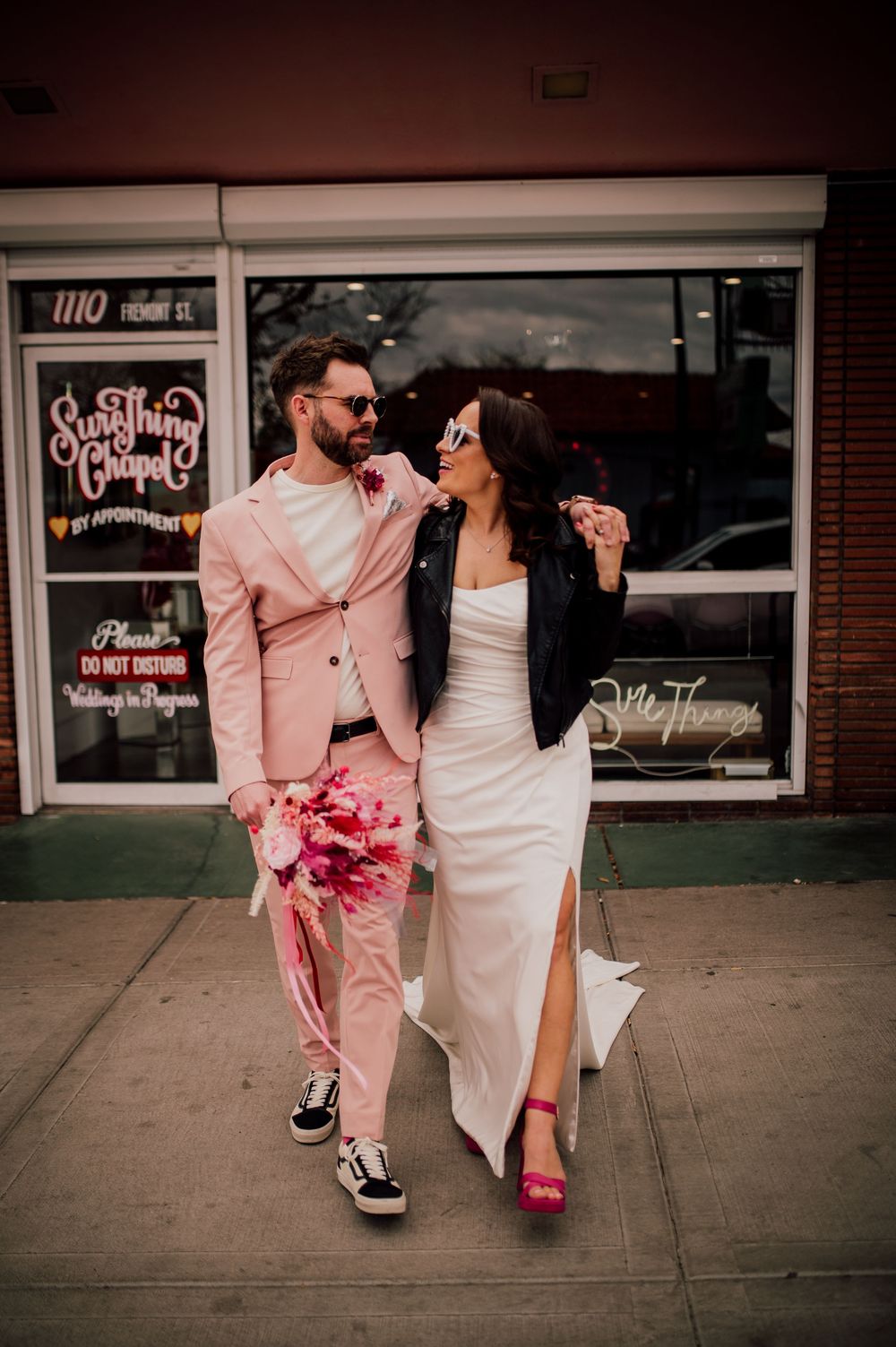 A couple in pink and white outfits walking together on a sidewalk holding hands and carrying pink flowers.