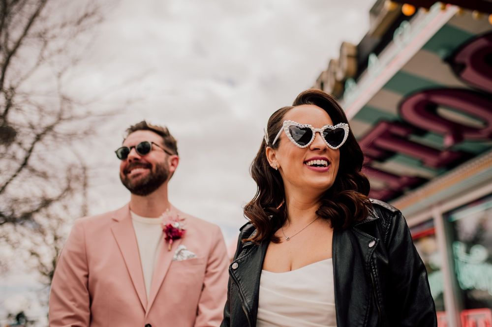 Stylish couple wearing sunglasses smile together outdoors, with one in pink suit and one in leather jacket.