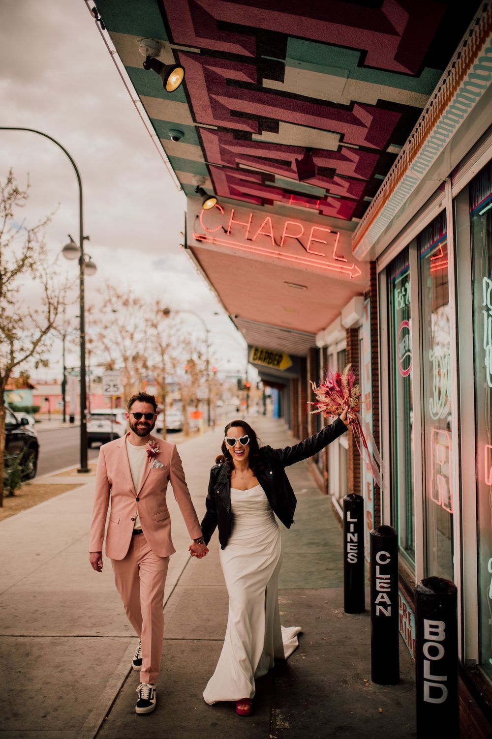A couple celebrates joyfully while walking under a vintage storefront sign on an urban street at sunset.