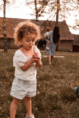 Curly-haired toddler in white outfit blowing a dandelion in a sunny field with women and rustic buildings behind.