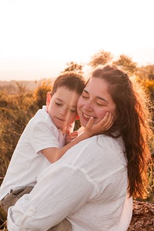 A mother and son share a tender embrace outdoors at golden hour, both smiling with eyes closed in a warm sunset glow.