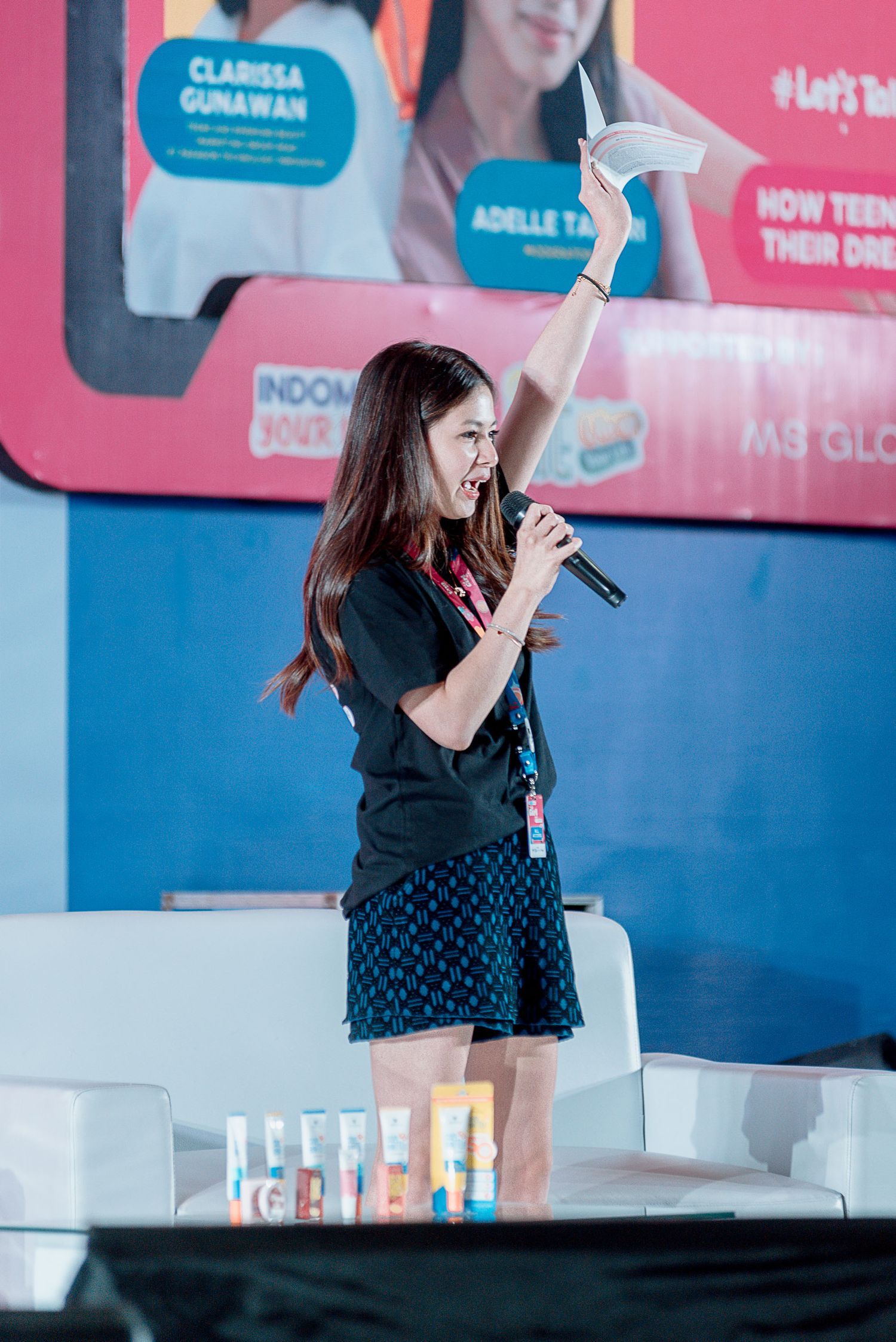 A performer in a black t-shirt and patterned skirt sings into a microphone while standing on stage with a blue backdrop.
