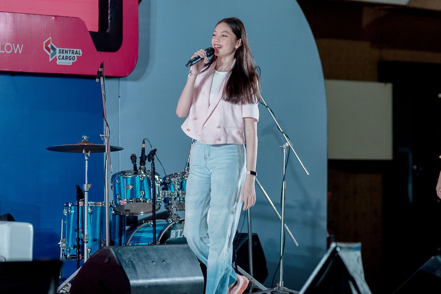 A performer in light blue pants and a pink top sings into a microphone on stage with blue lighting.