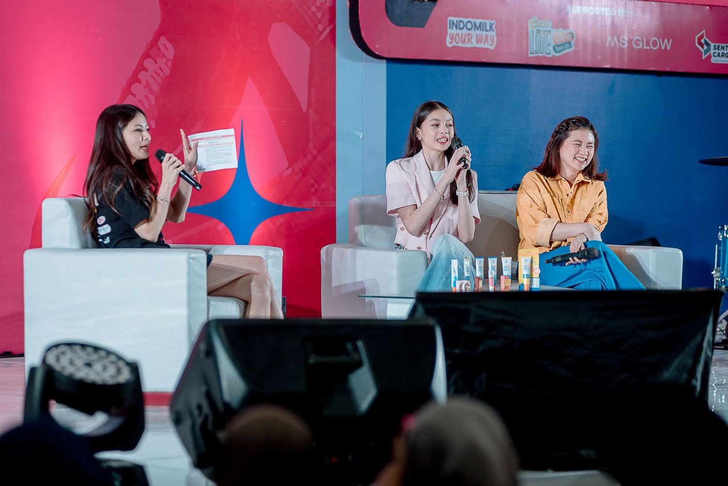 Three people sit on a panel stage with pink and blue backdrop lighting during a discussion event.
