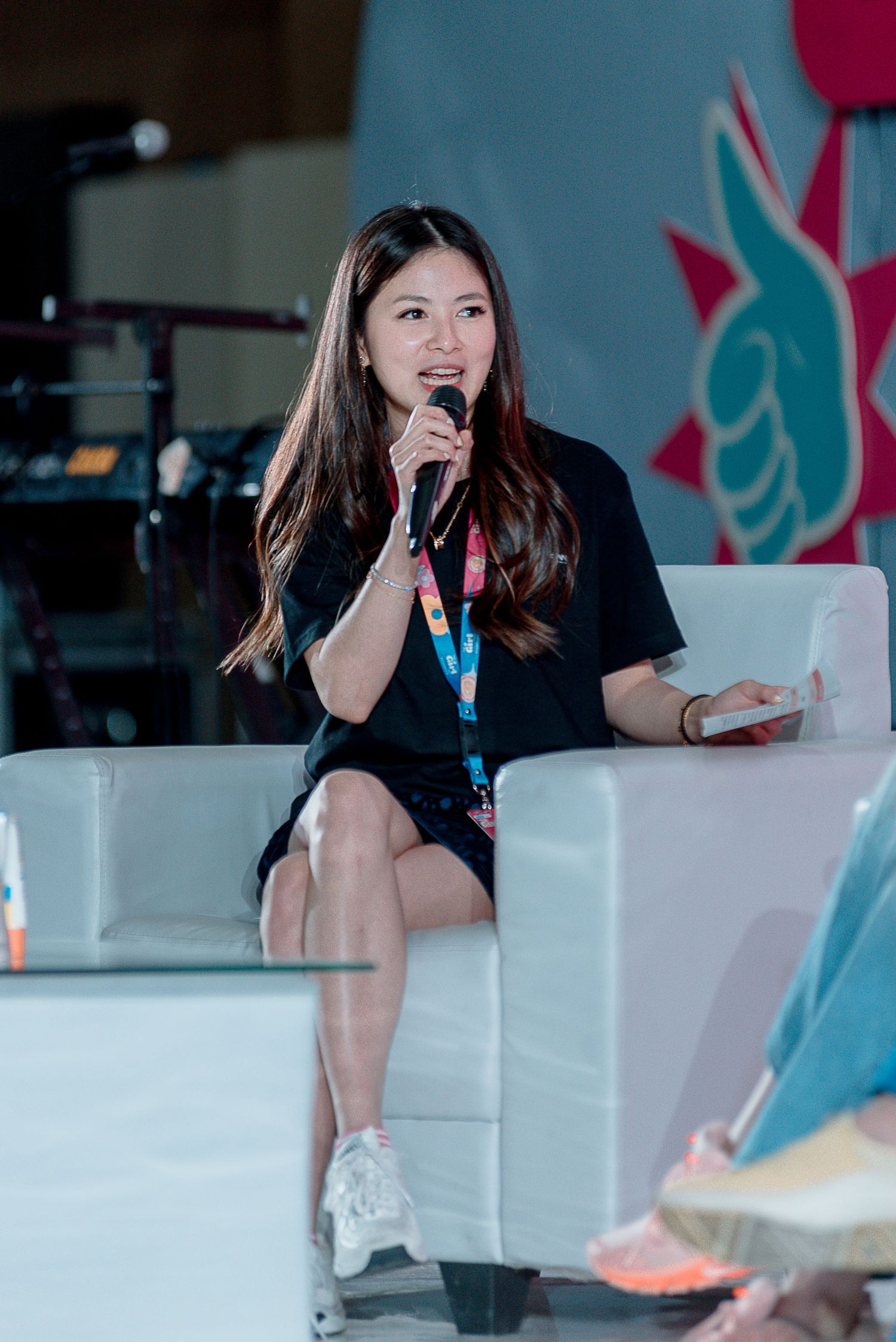 A speaker in a black shirt sits in a white chair on stage during a presentation at a conference.