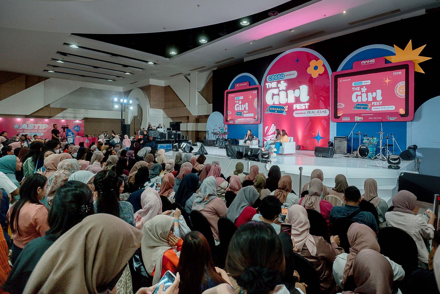A crowded conference room filled with attendees watching presentations on large screens displaying pink branding elements.