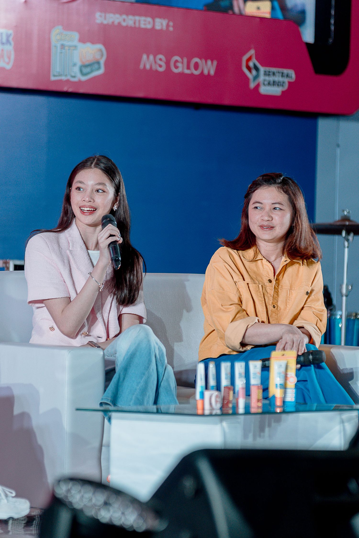 Two people sit on a couch at an event with MS Glow branding visible in the background.