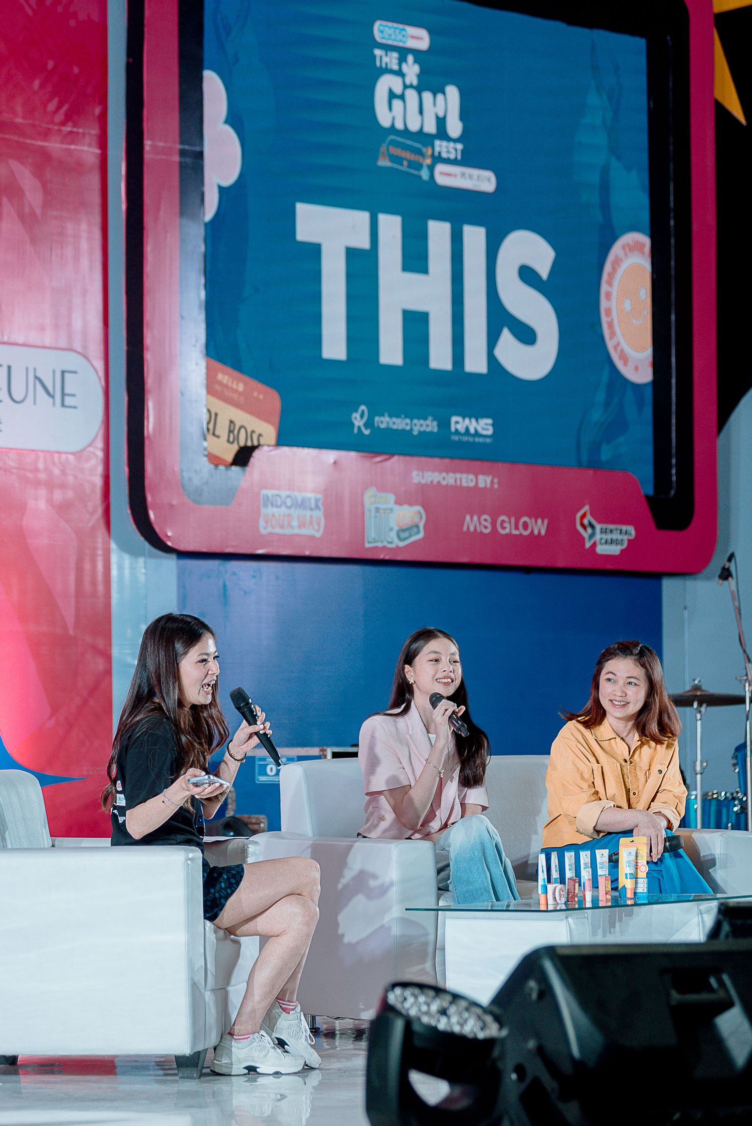 Three speakers sit on stage at a panel discussion event with blue and pink lighting and signage.