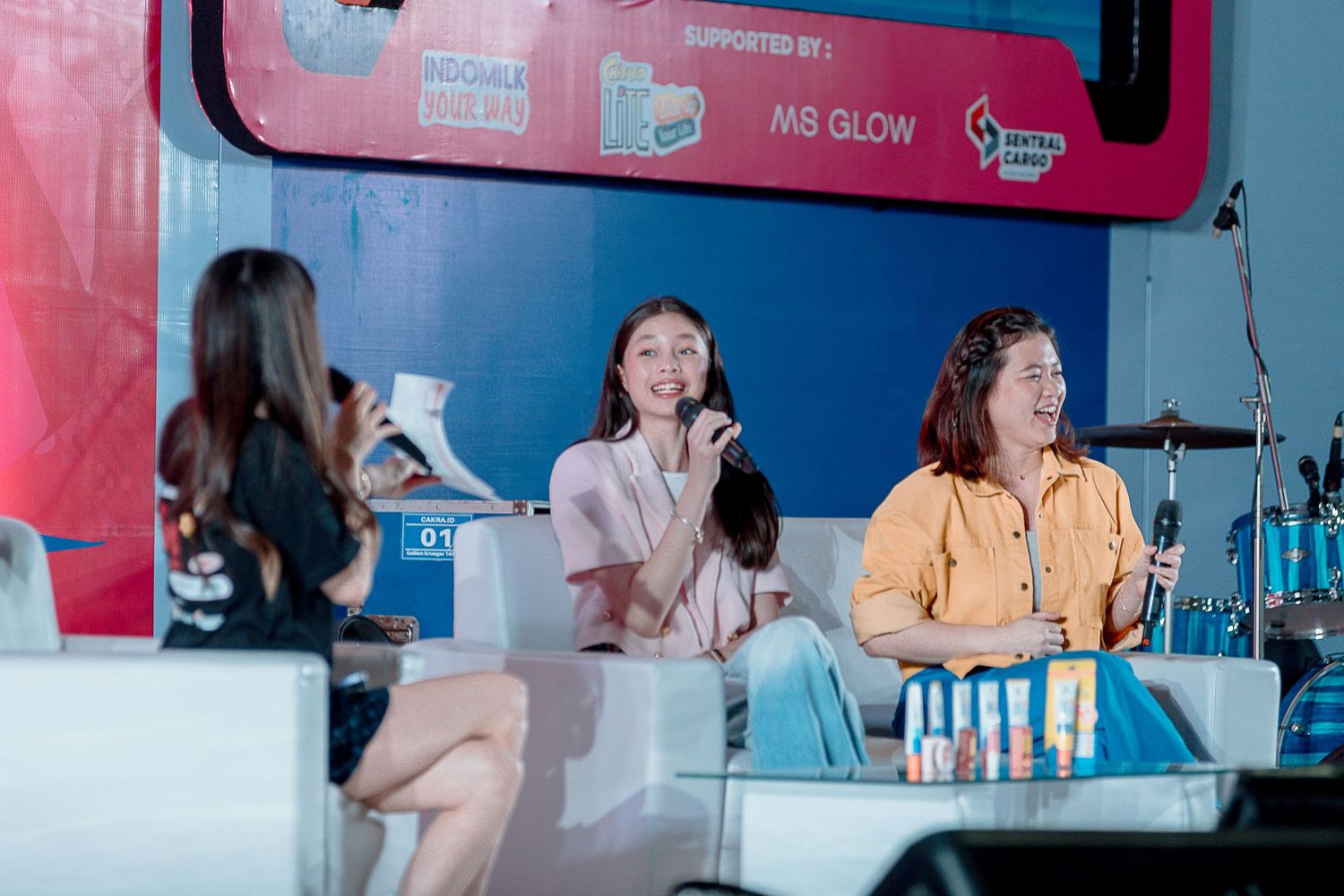 Three people sit at a pink-themed promotional event booth with product displays and microphones.