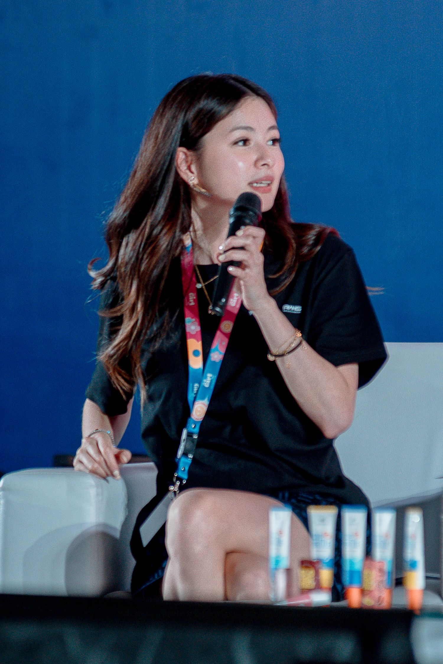 Professional speaker in black attire gestures while presenting at a conference with blue backdrop.