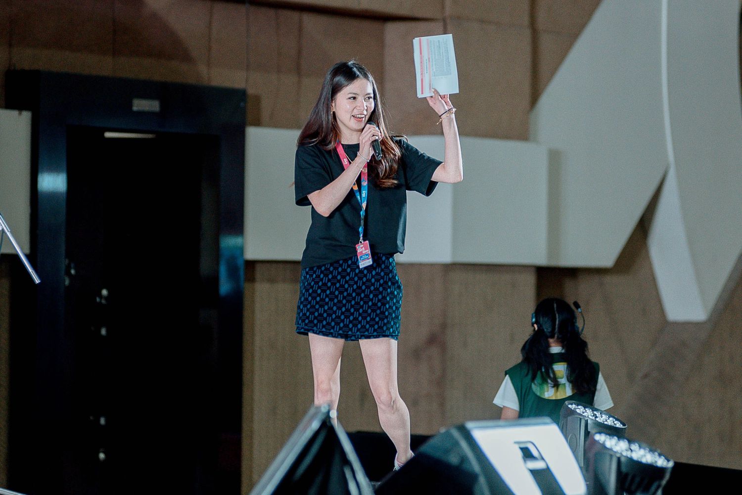 A speaker on stage holds up papers while presenting at a conference in a polka dot dress and microphone.
